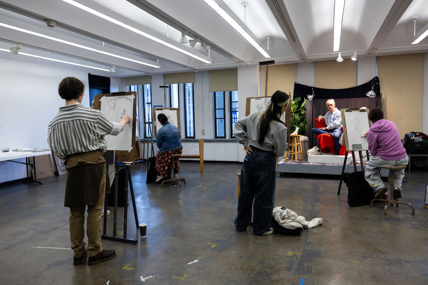 Four students stand at easels and sketch a live model who is seated at the front of the class in front of a red cloth backdrop.