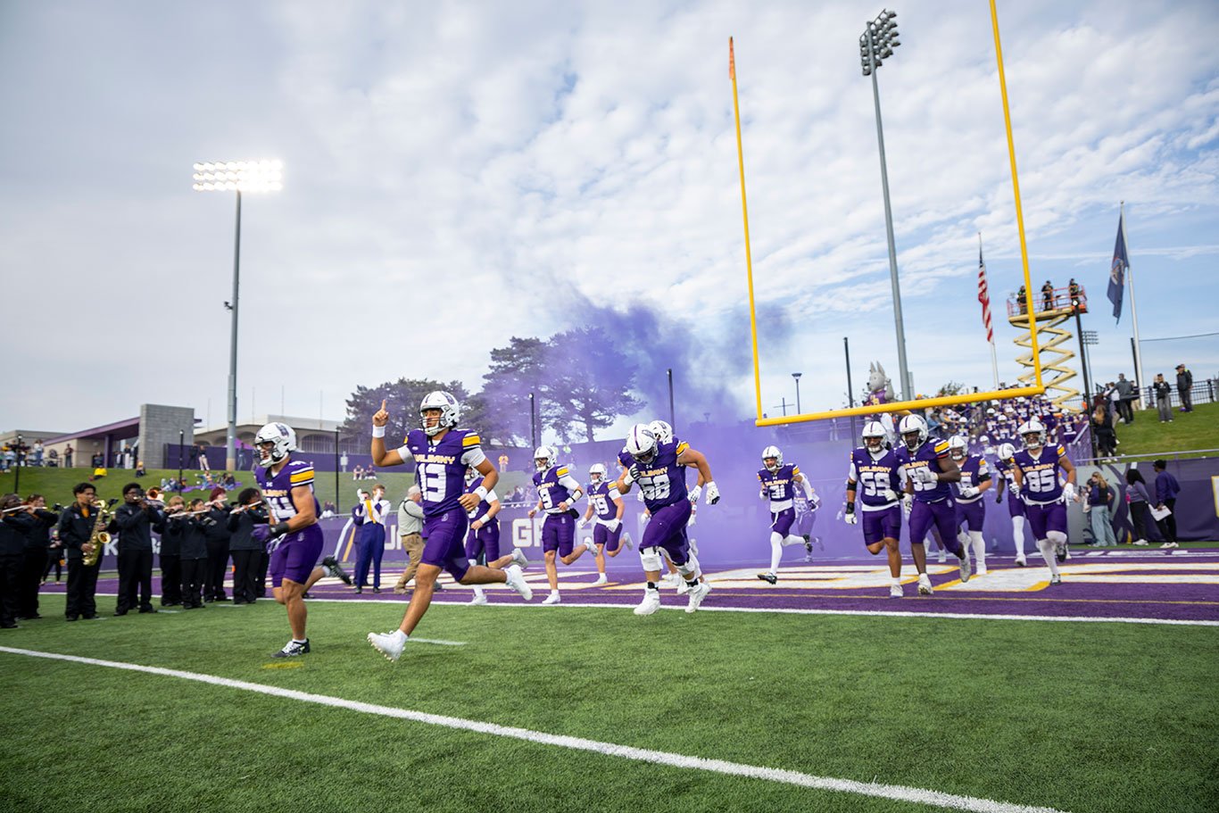 UAlbany football players running through purple smoke on to the field for the 2025 Homecoming game.
