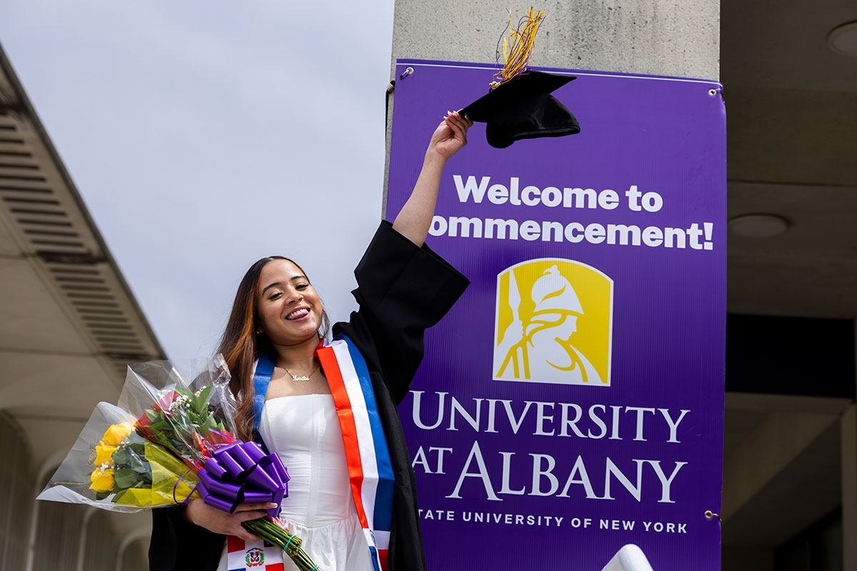 Female student in a white dress holding her mortar board up high in front of a Welcome to Commencement sign.