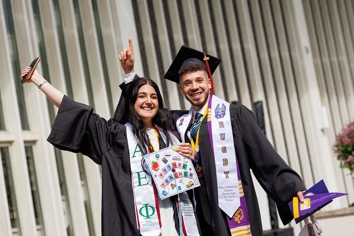 Two students in full regalia posing with their arms in the air.