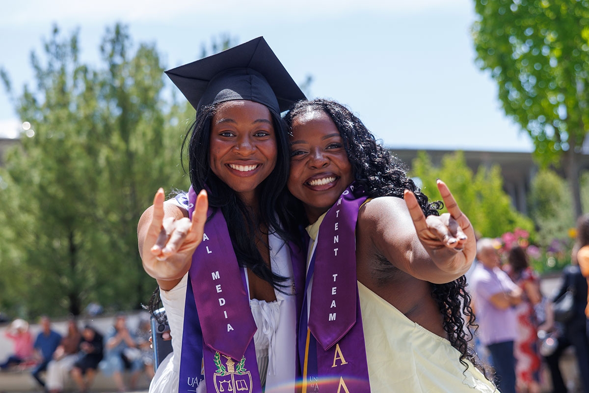 Two female students in regalia smiling and making Damien with their hands.