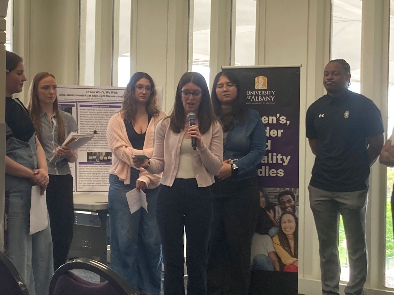 Five students standing behind a student talking on a microphone.