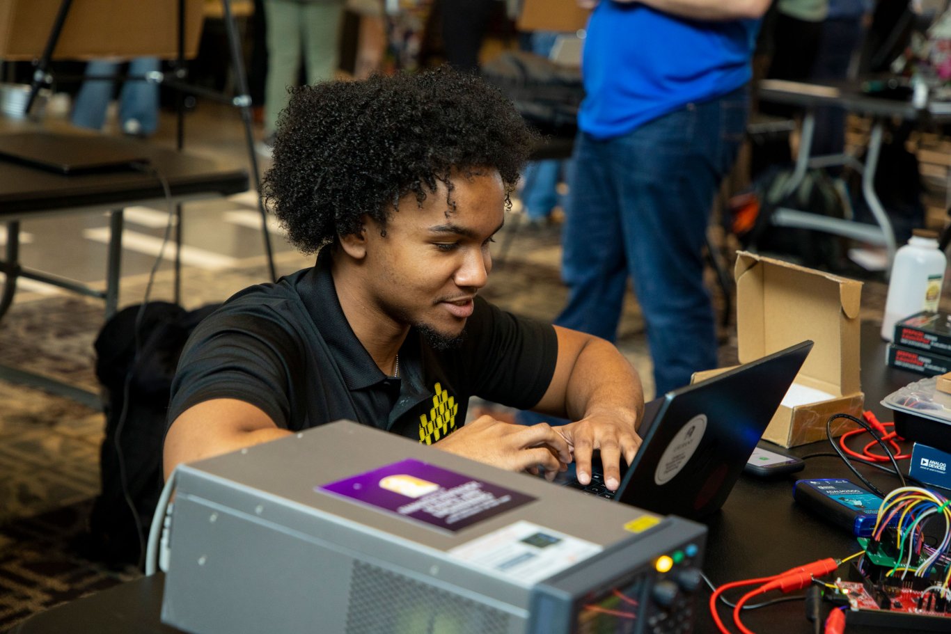 An engineering student crouches to work on a laptop placed on a table where wires connect to a CPU.