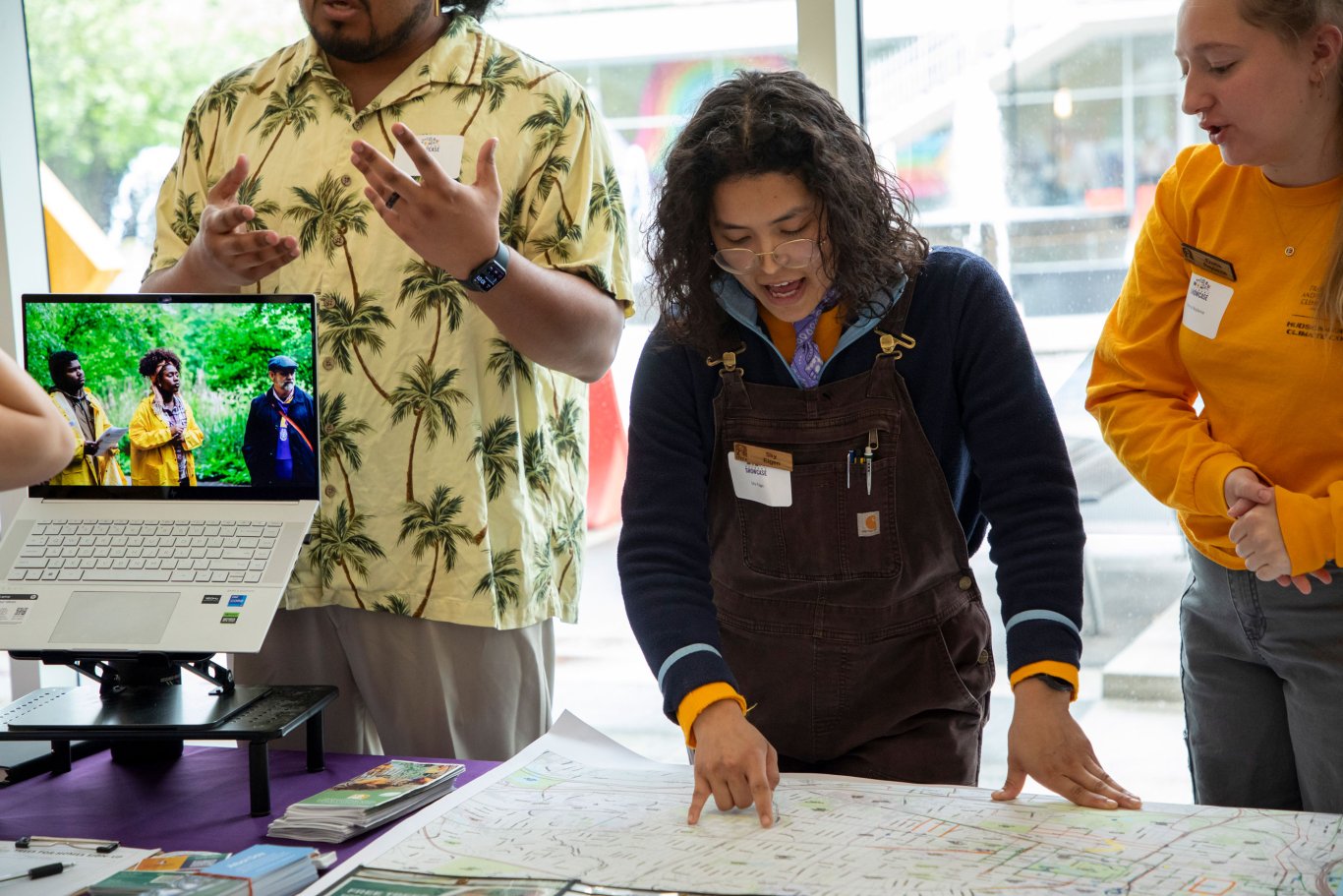 Three student presenters stand behind a table and speak with Showcase attendees. Two students gesture towards a map, while the third gestures towards a slideshow playing on a laptop.