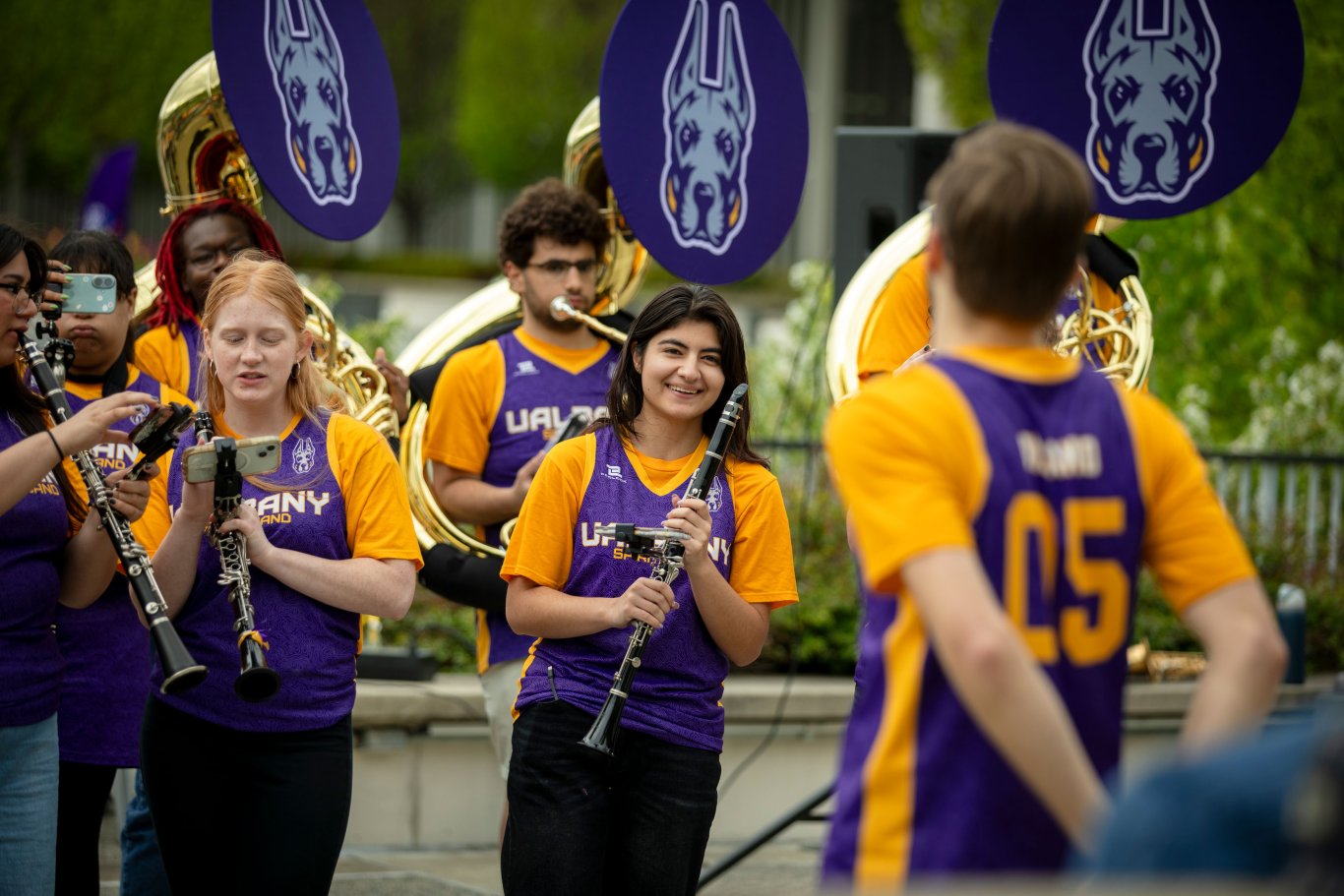 A clarinet player smiles at the conductor while surrounded by fellow UAlbany Spirit Band members, who are all wearing matching purple and gold jerseys.