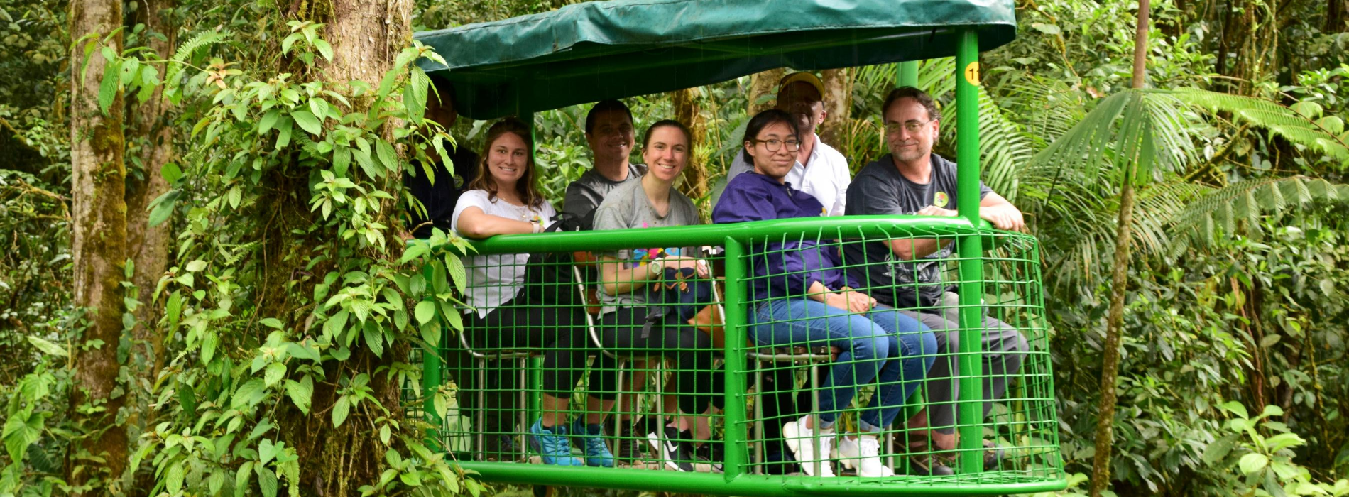 A group of students and faculty sit on a traveling cart in the rainforest. They are smiling at the camera.