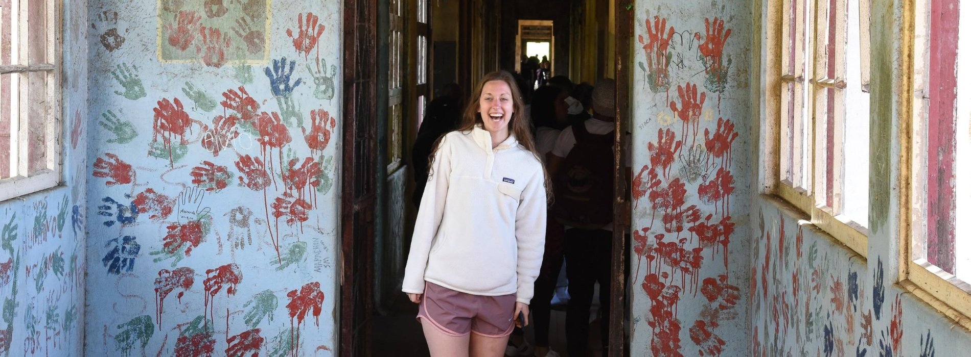 A student smiles brightly at the camera, standing in a building painted with handprints on a study abroad trip. 