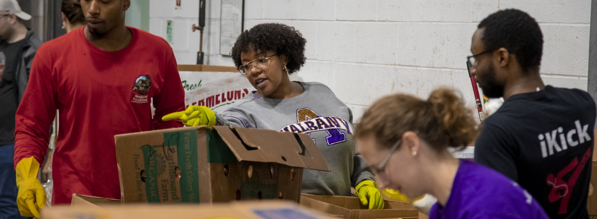 A student at the food bank points at a box of food that needs to be sorted. She is standing behind several boxes of packaged food.