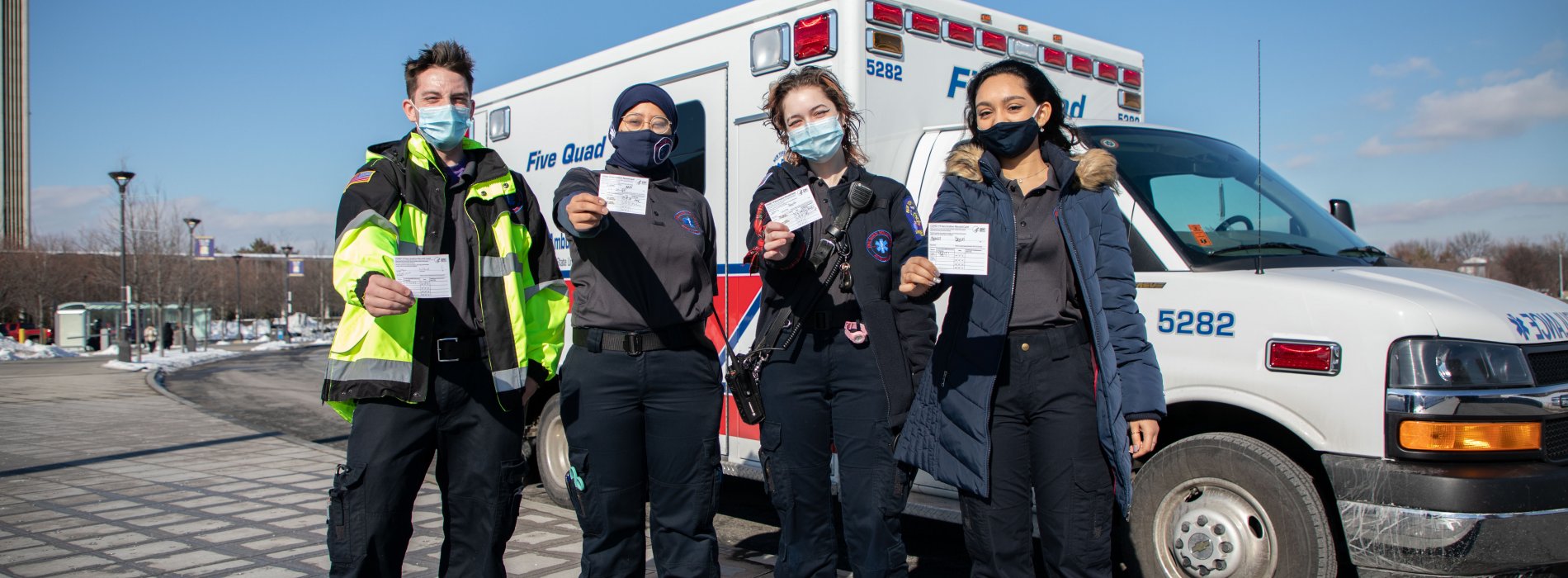 Four student EMTs pose in front of the Five Quad ambulance with their COVID-19 vaccination cards