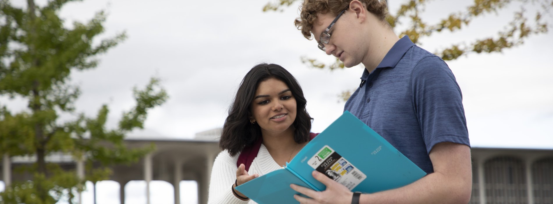A student holds a notebook to study with classmate outside on campus