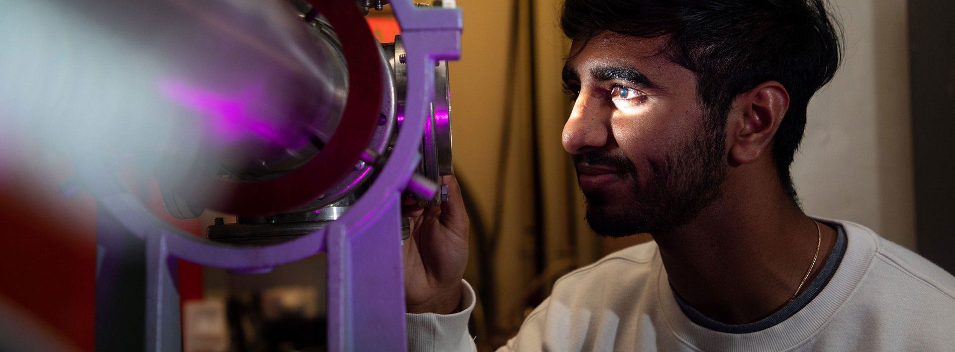 A researcher smiles as he looks into a piece of equipment lit up in purple inside the Ion Beam Lab.