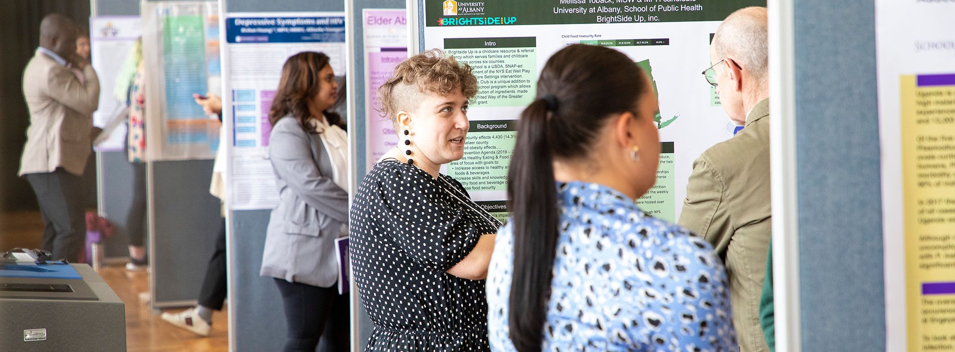A line of academic posters is set up in an auditorium. Students stand in front of their posters, presenting to guests.
