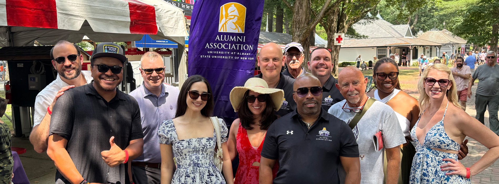 A group of UAlbany alumni stand in a group by the festival tent at the Saratoga Race Course.