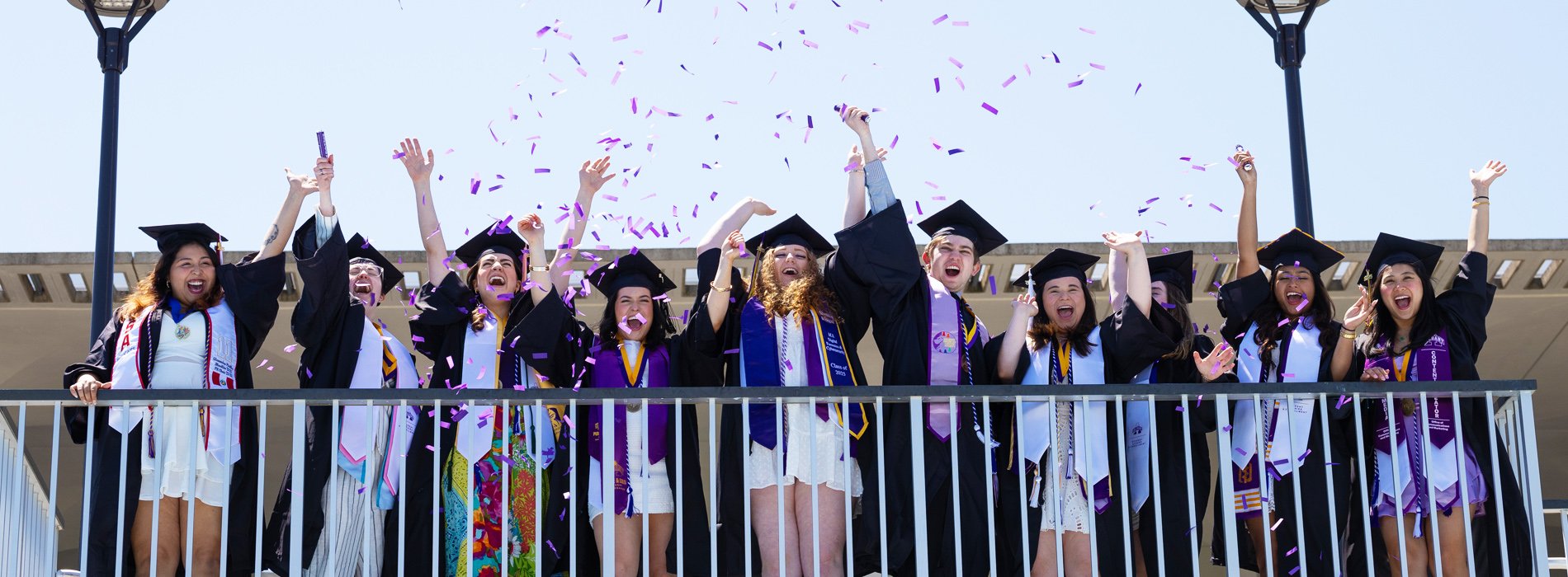 A group of graduates stand behind a fence and throw purple confetti in the air.