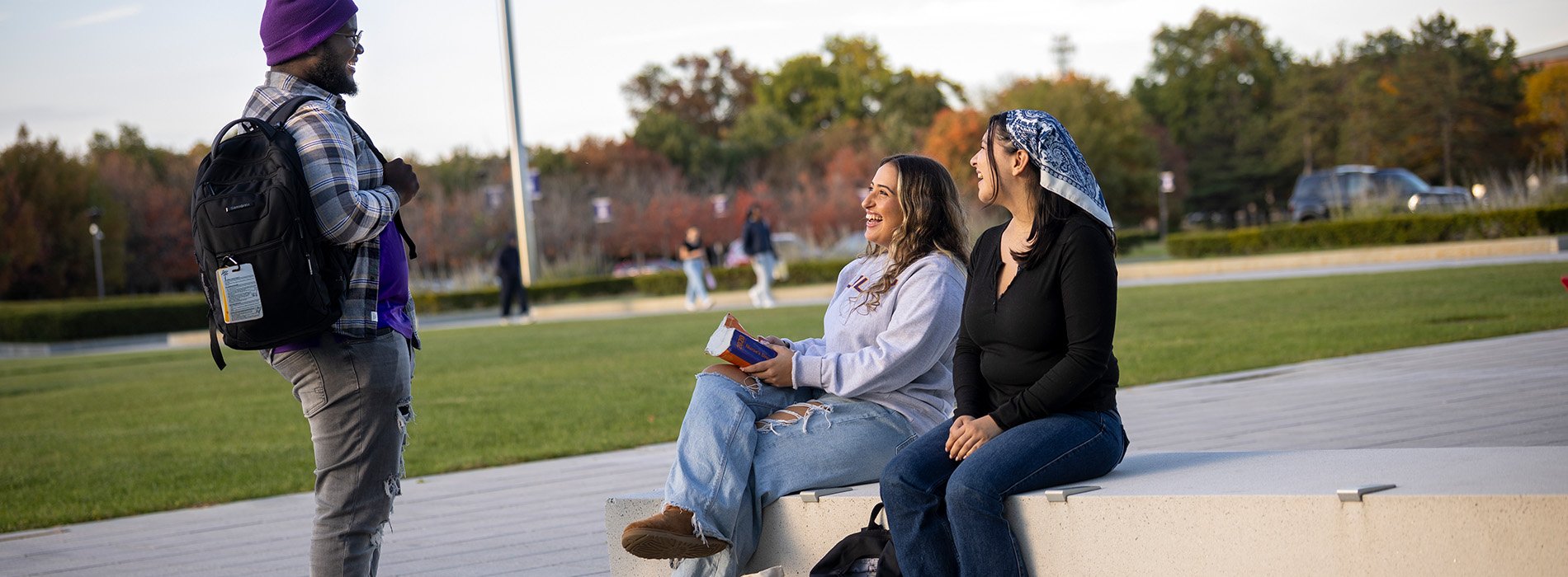 A group of three students interact on campus. One is standing and two are sitting as they talk and laugh with each other.