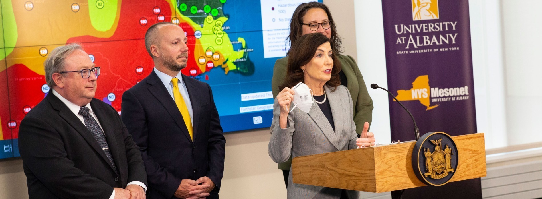 New York State Governor Kathy Hochul holds up a face mask and speaks at a press conference while standing in front of a NYS Mesonet map and UAlbany logo.