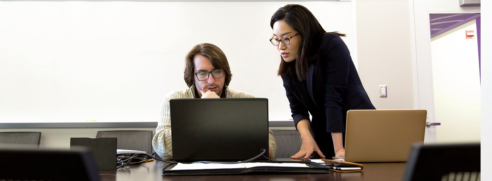 A woman in glasses and a black blazer stands a looks over the shoulder of a student in a tan sweater and glasses. Both look at a laptop on a table.