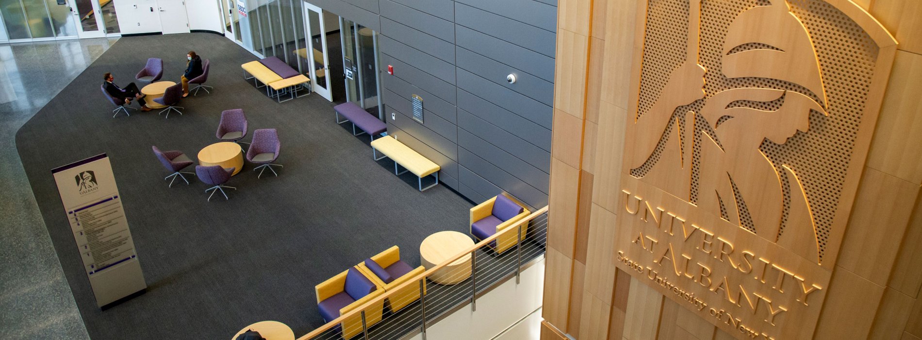 Two people sit in purple chairs inside the ETEC lobby, which has a large carved wood sign that says University at Albany, State University of New York.