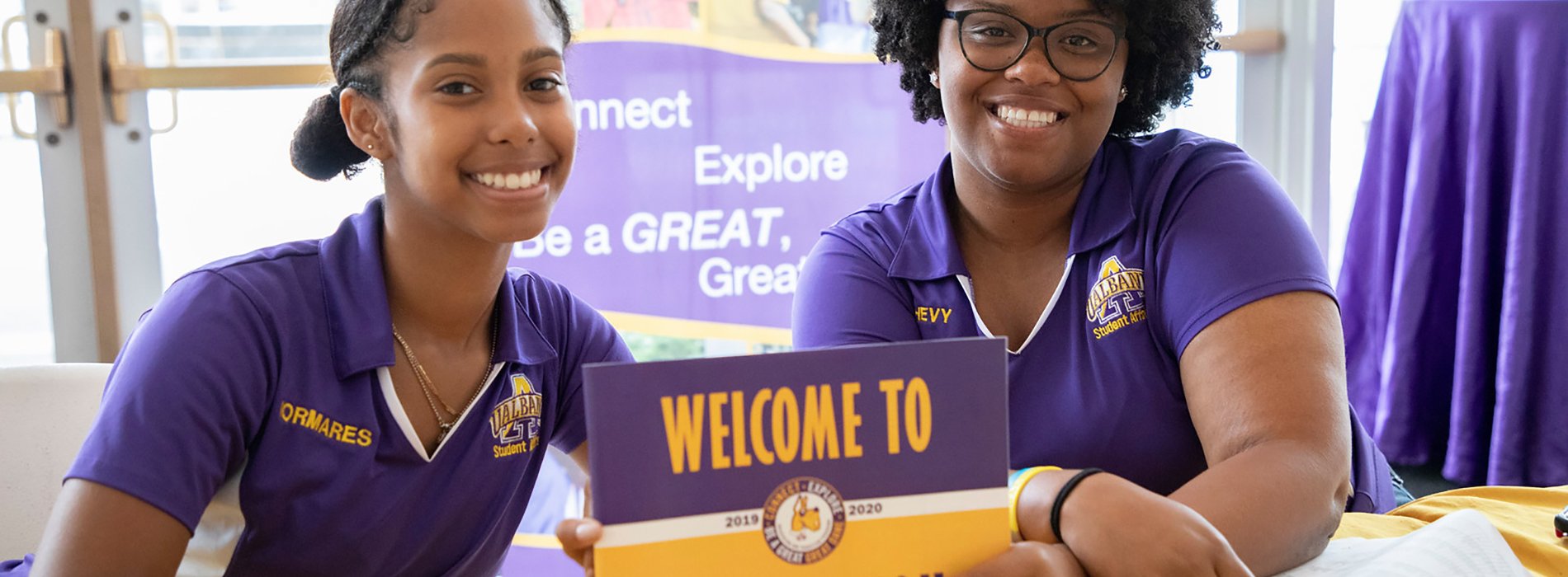 Two Orientation Leaders pose for a photo at a welcome desk, while holding a "Welcome to Orientation" sign.