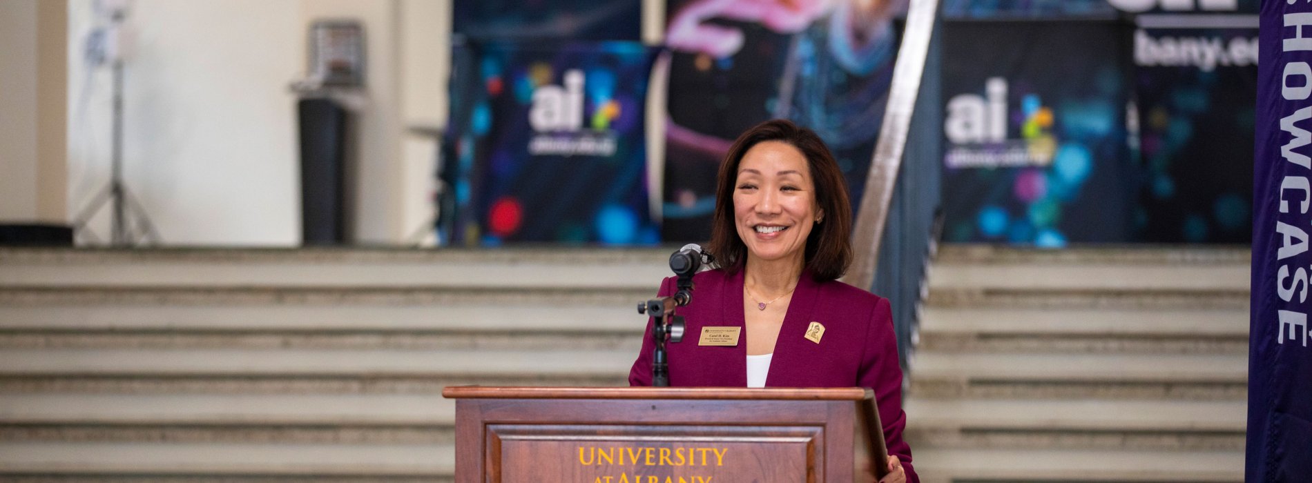 UAlbany Provost Carol Kim smiles while speaking at a University podium, with posters for the AI-Plus initiative behind her.