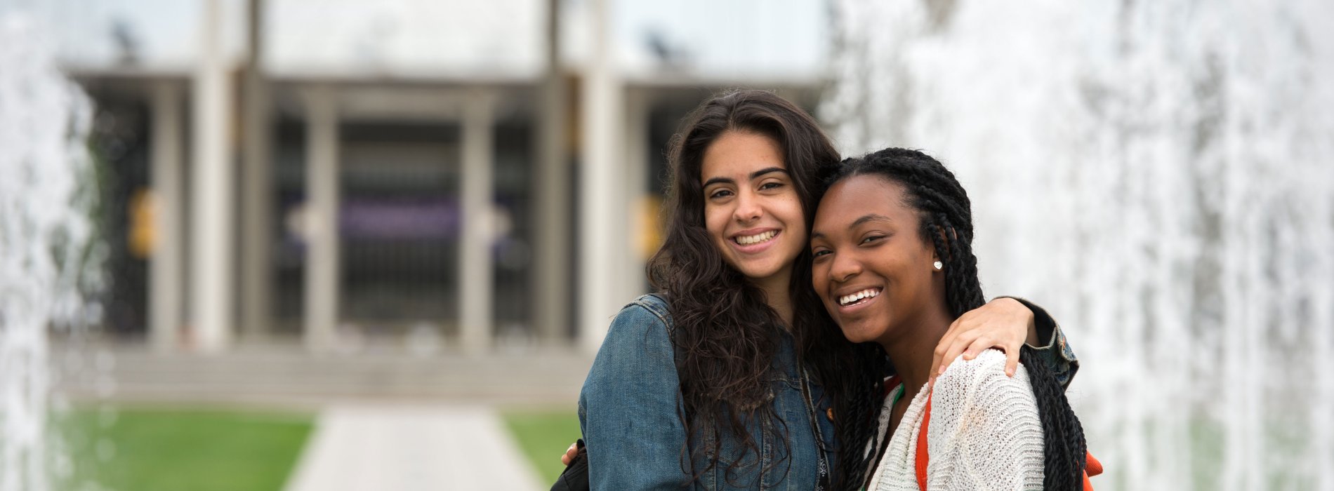 Two students with backpacks embrace as they smile and pose for a photo in front of fountains on the Uptown Campus.