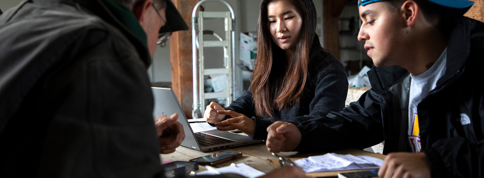 Two students sit inside a barn at a table with the farm owner reviewing notes.