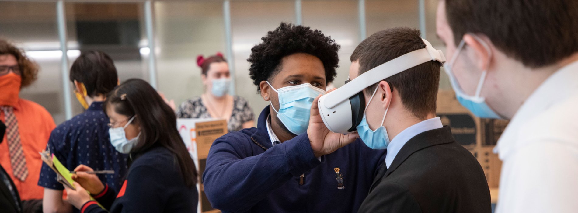 A student helps another person put on a virtual reality head seat during a poster session presentation.