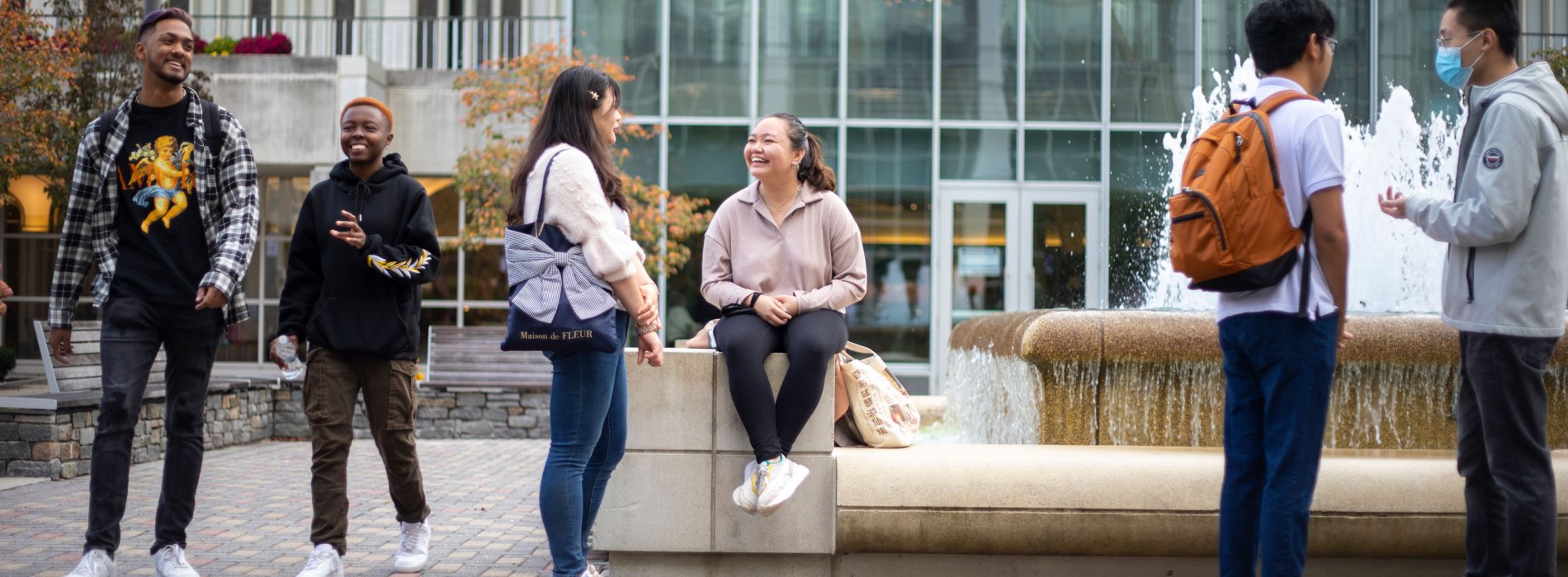 Six students gather outside on campus, speaking and smiling in pairs. Two are walking, three are standing and one is sitting.