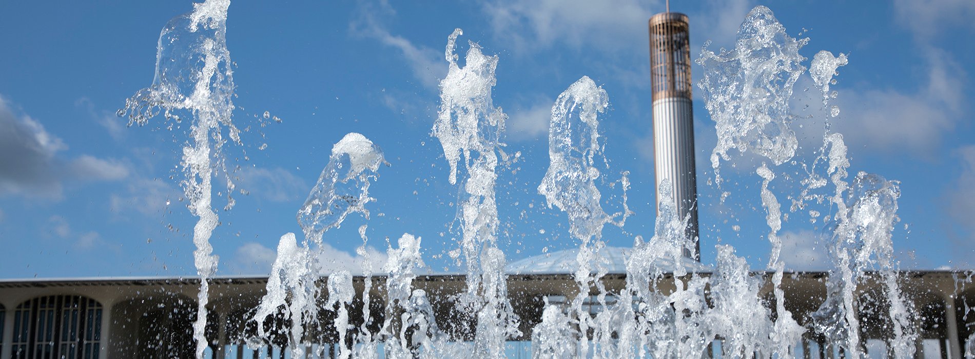 The UAlbany carillon and Academic Podium are seen behind a fountain, under blue skies