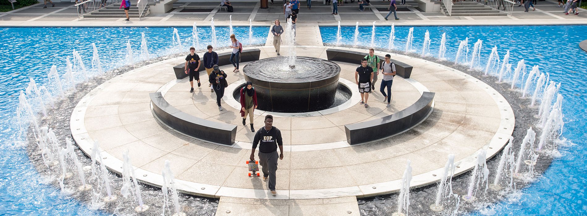 Students walk through UAlbany's academic podium, which includes staircases, overhangs, benches and a fountain.