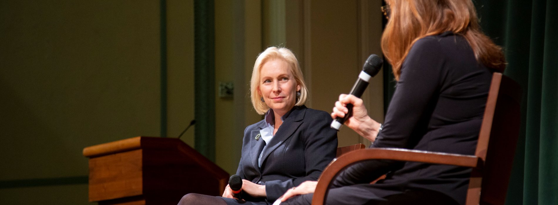 U.S. Senator Kirsten E. Gillibrand sits on stage holding a microphone as she is interviewed by another woman.