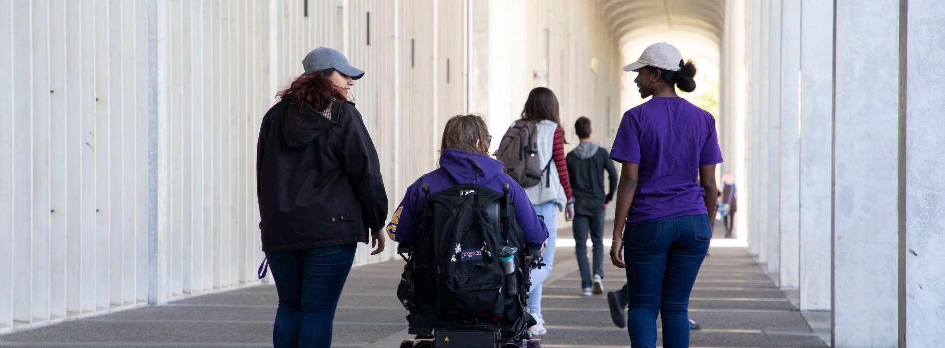 Two students walk on either side of a third student using a motorized wheelchair under the arches of UAlbany's Uptown Campus.