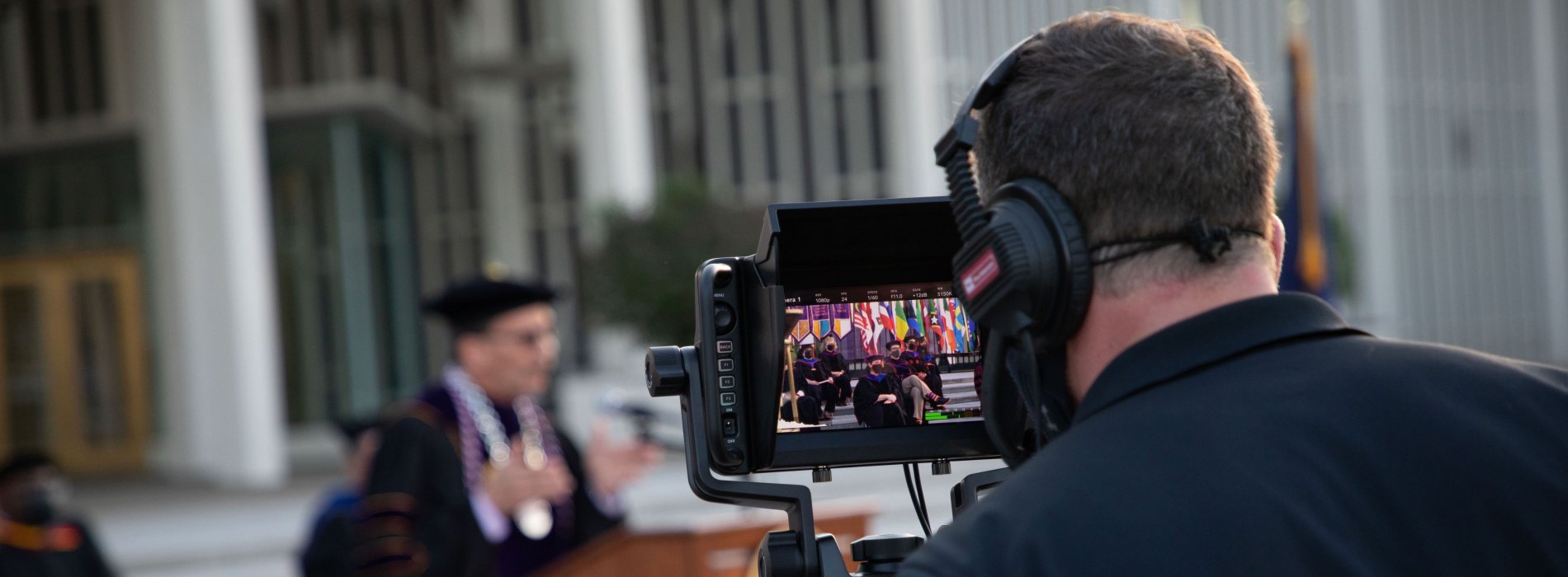 A camera operator wearing headphones looks into a camera display screen as he records Commencement