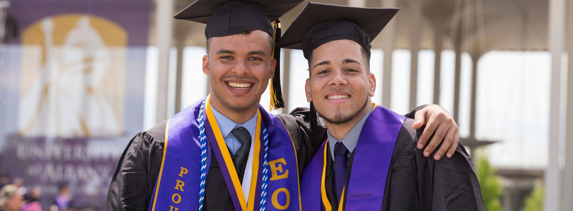 Two students wearing graduation caps, gowns and stoles pose for a photo. One graduate is wearing a Proud to Be EOP stole.