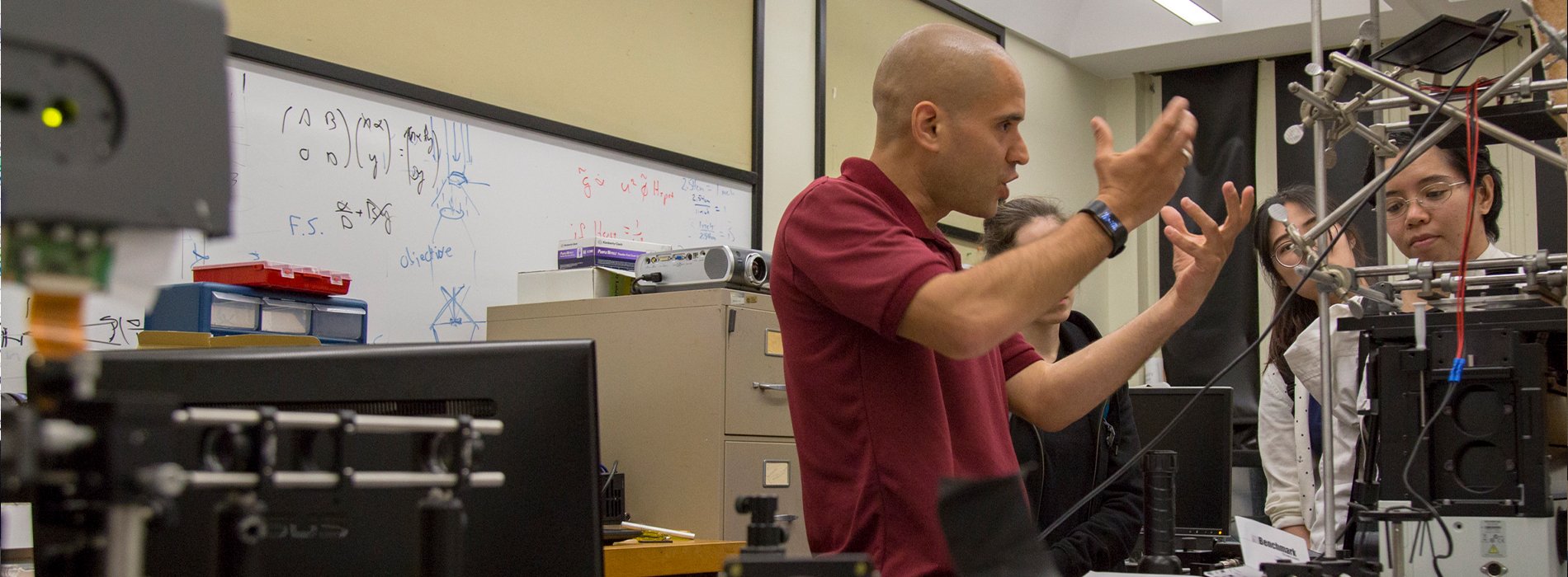 A physics instructor explains a concept to three students while standing in a lab, surrounded by physics equipment and a white board with equations.