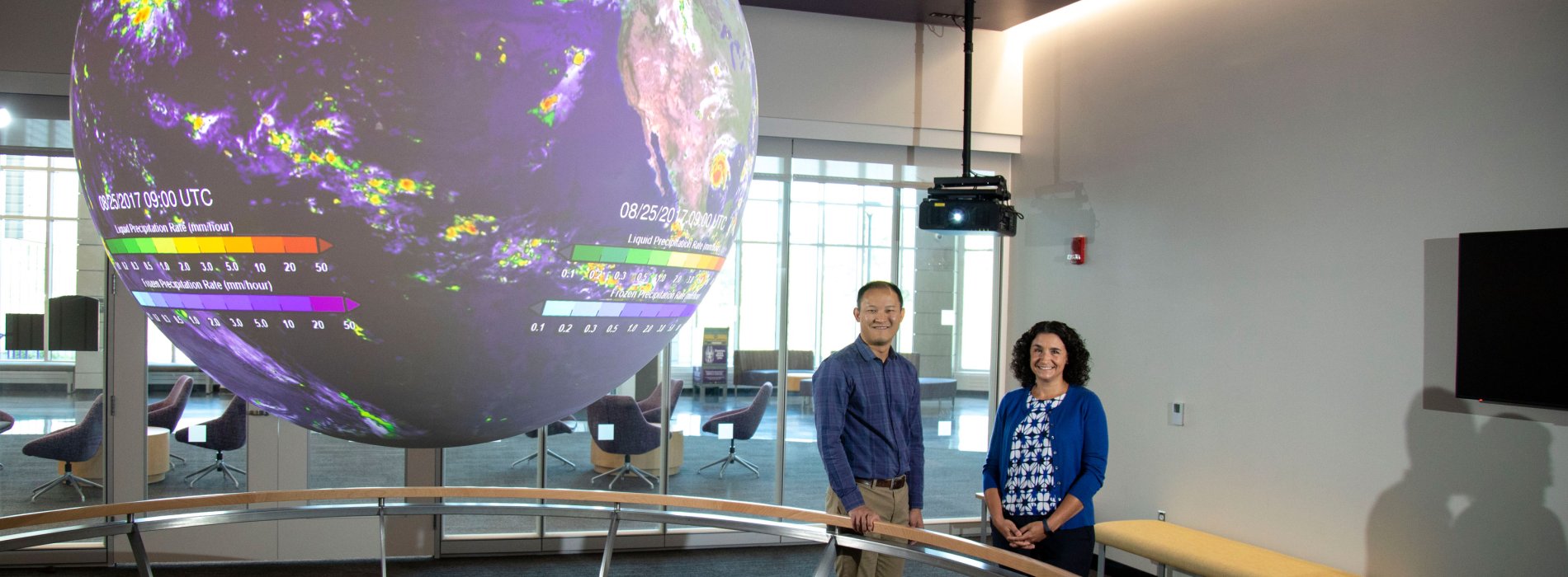 A man and a woman stand and smile for a camera in front of a huge digital globe showing live weather patterns.