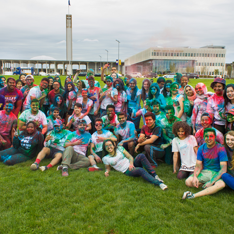 A large group of students pose for a photo after participating in Holi. They are all covering in varying degrees of colorful powder, some throw the powder up into the air in celebration.