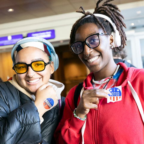 Two UAlbany students pose and smile, each holding up a sticker that reads "I Voted."