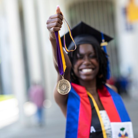 A student wearing a graduation mortar board smiles and holds up an honors medallion.