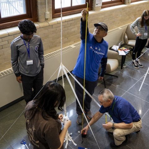 As two students set up a tower, an instructor squats down to hold a tape measure while a volunteer pulls it up.