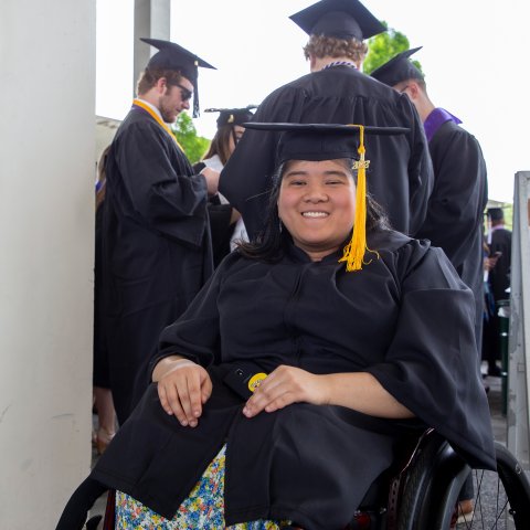 A student smiles during commencement, wearing a cap and gown. They are sitting in a wheelchair with a small group of others behind them.