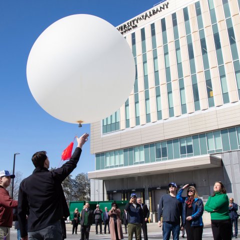 A man releases a large white weather balloon in front of about a dozen people standing outside ETEC at UAlbany.