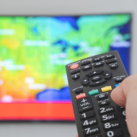 Hand holding a remote control in front of a TV displaying weather map.