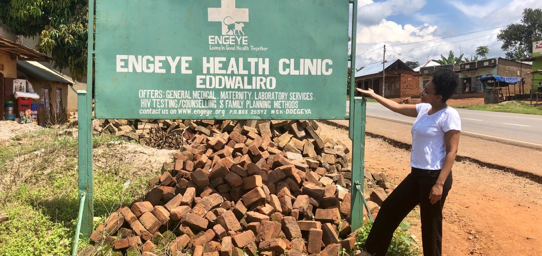 Elizabeth S. stands next to a green sign in Uganda that says "Engeye Health Clinic".