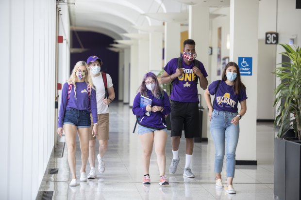 Five students wearing UAlbany shirts and masks walk through a hallway on campus