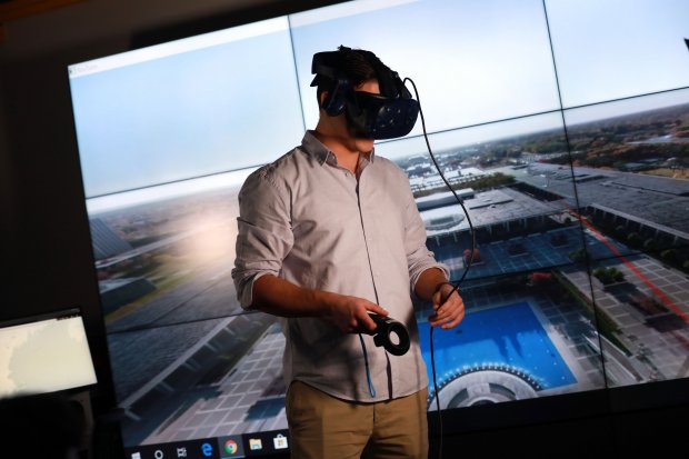 A researcher wears a virtual reality headset while standing in front of a large screen inside the xCITE lab.