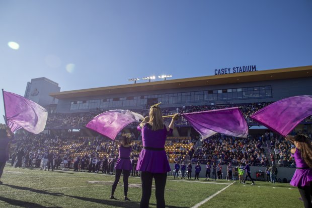 Color guard performs with purple flags on turf football field in Casey Stadium before hundreds of fans