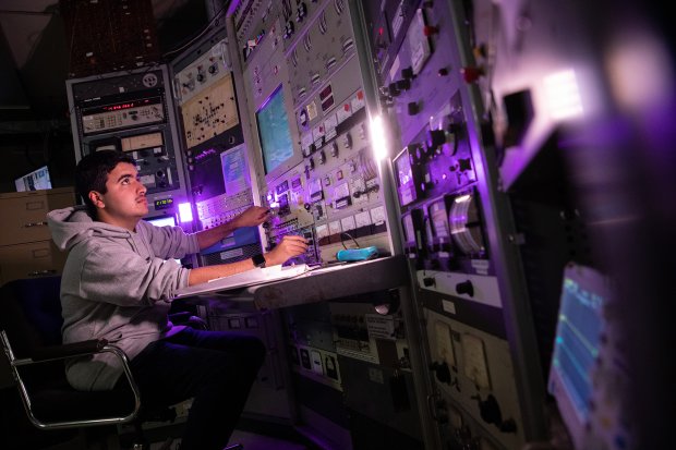 A student sits at a desk in front of a large instrument board and turns a different dial with each hand, as he looks up at the monitor.