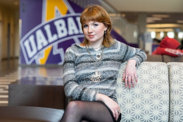 A woman with short red hair and a lip ring wears a gray and white fuzzy sweater over a skirt and tights with chunky necklaces. She sits on a cushioned seat in the Campus Center, one arm folded over her lap.