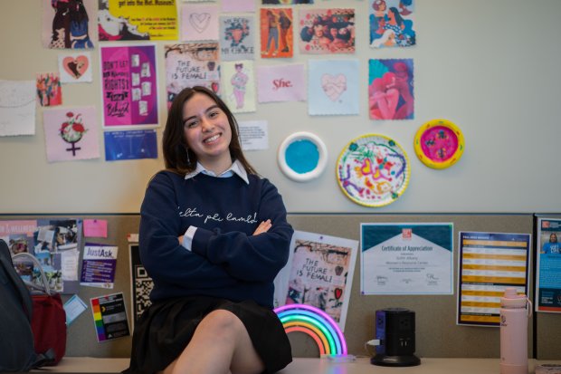 A woman with long brown hair wears a navy sweater over a collared shirt and black skirt and poses seated with her arms crossed. In the background colorful posters and art dot the wall.